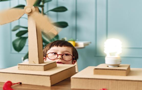 Junge vor Papierwindkraftwerk mit leuchtender Glühbirne, (c)iStock.com/pinstock